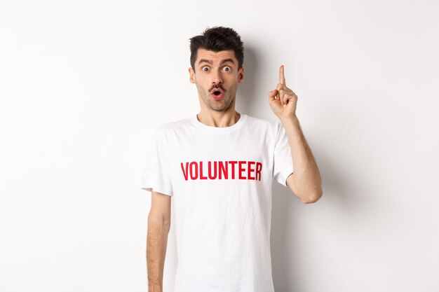 Apuesto joven en camiseta de voluntario teniendo una idea, levantando el dedo y diciendo sugerencia, apuntando hacia arriba, de pie sobre fondo blanco.