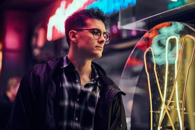 Un apuesto joven con capucha deportiva parado en la calle por la noche. Letreros iluminados, neón, luces.