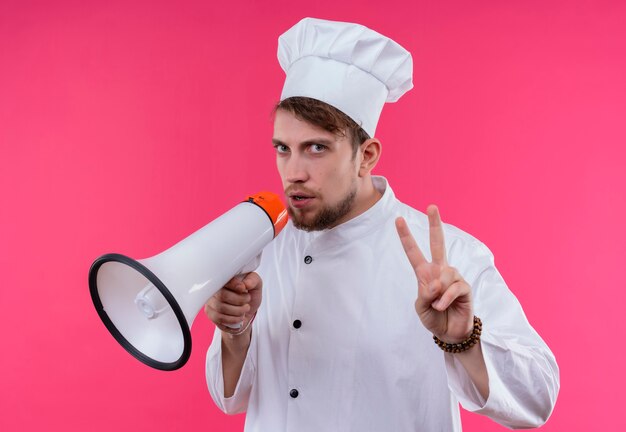 Un apuesto joven chef barbudo con uniforme blanco hablando por megáfono y mostrando dos dedos mientras mira en una pared rosa