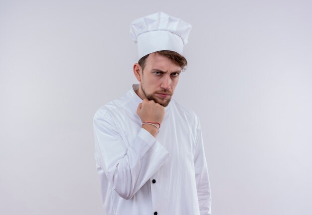 Un apuesto joven chef barbudo vestido con uniforme de cocina blanco y sombrero mostrando el puño cerrado mientras mira con expresión agresiva en una pared blanca