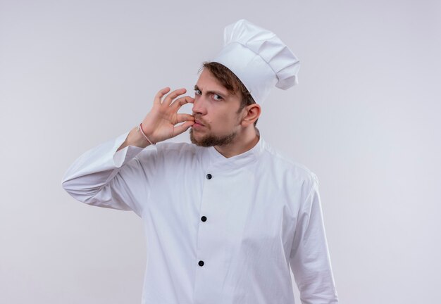 Un apuesto joven chef barbudo vestido con uniforme de cocina blanco y un sombrero que muestra un sabroso signo de ok con los dedos mientras mira seriamente en una pared blanca