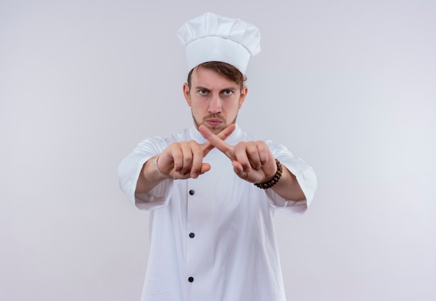 Un apuesto joven chef barbudo vestido con uniforme de cocina blanco y sombrero que muestra el signo x con los dedos mientras mira con expresión agresiva en una pared blanca