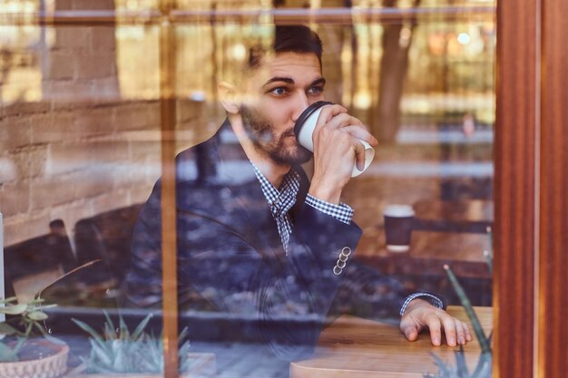 Un apuesto joven elegante con traje clásico bebiendo café en el café detrás de la ventana.