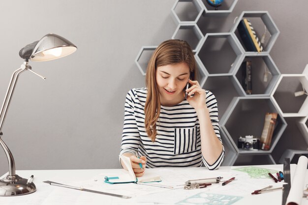 Apuesto joven emprendedor diseñador con cabello oscuro en camisa a rayas hablando por teléfono con el cliente discutiendo detalles del diseño del sitio web y escribiéndolos en el cuaderno antes de reunirse en la vida real