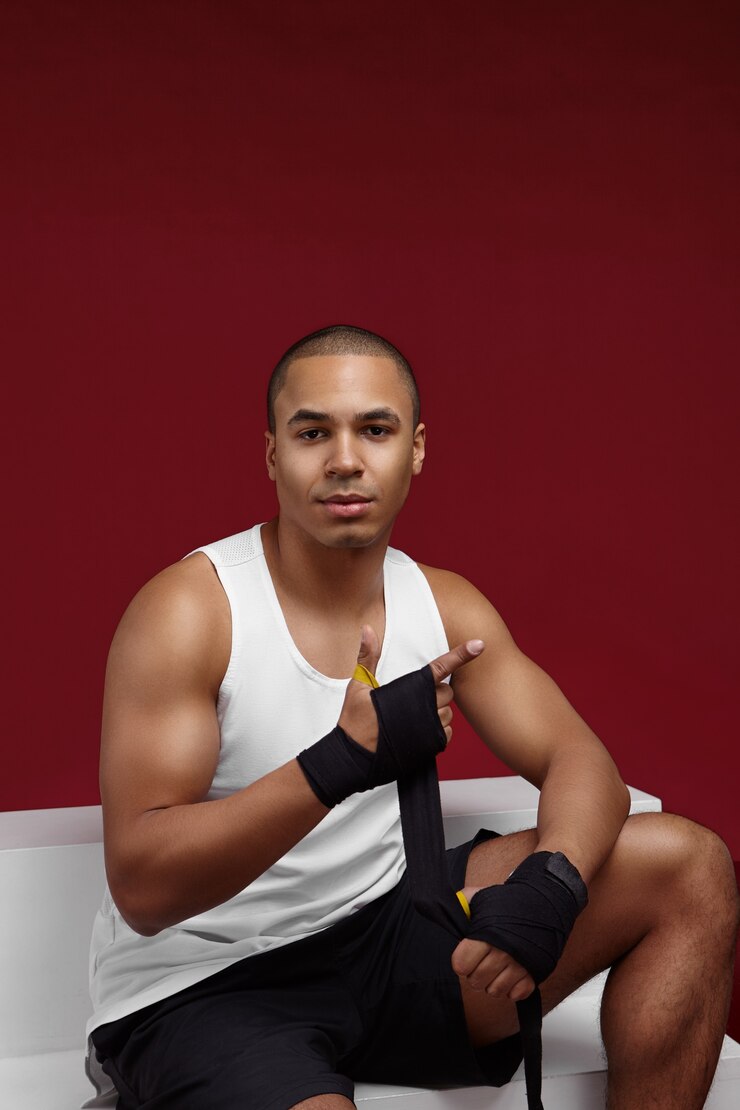 Foto gratuito apuesto joven empresario africano confiado con hombros musculosos con pantalones cortos negros y camiseta sin mangas blanca atando vendas de boxeo para entrenar con saco de boxeo, entrenando después del día de trabajo en la oficina