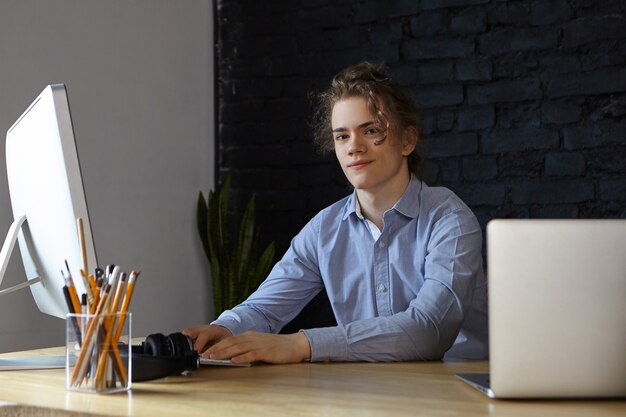 Apuesto joven empresario exitoso vestido con camisa azul sonriendo, desarrollando un nuevo proyecto empresarial de puesta en marcha en su lugar de trabajo, teniendo grandes ideas y planes, utilizando dispositivos electrónicos modernos