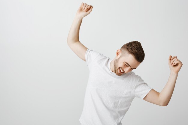 Apuesto joven feliz bailando en camiseta blanca sobre pared gris