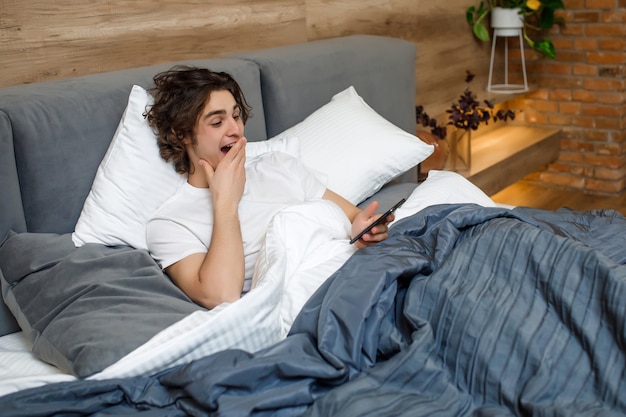 Apuesto joven feliz en pijama usando un teléfono celular mientras está acostado en la cama por la mañana