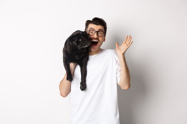 Apuesto joven con gafas sosteniendo su pug negro y saludando con la mano, chico saludando mientras lleva el perro con un brazo, de pie sobre fondo blanco.