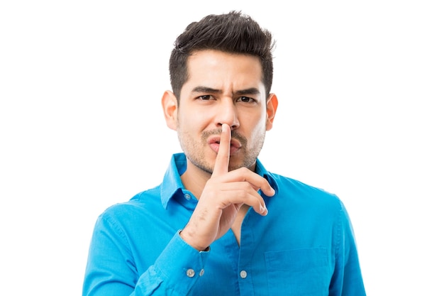 Apuesto joven gesticulando silencio con el dedo en los labios sobre fondo blanco.