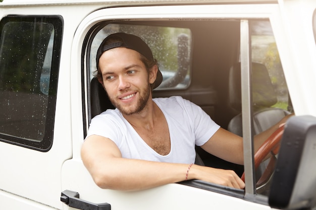 Apuesto joven con gorra de béisbol hacia atrás conduciendo un vehículo SUV blanco, sacando el codo de la ventana abierta, sonriendo