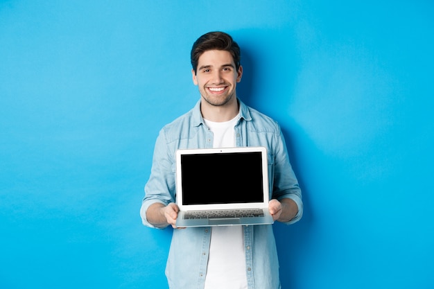 Apuesto joven introducir el producto en la pantalla del portátil, mostrando la computadora y sonriendo, de pie sobre fondo azul.