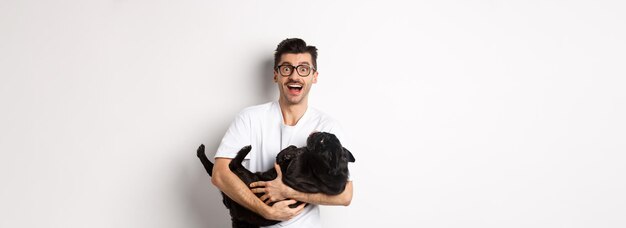 Apuesto joven jugando con el dueño del perro cachorro negro lindo acariciando a un pug de pie sobre backgro blanco