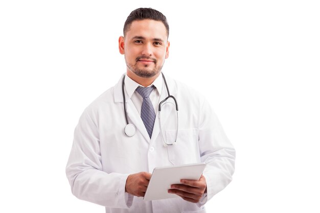 Apuesto joven médico con bata de laboratorio y estetoscopio usando una tableta para verificar el historial de un paciente