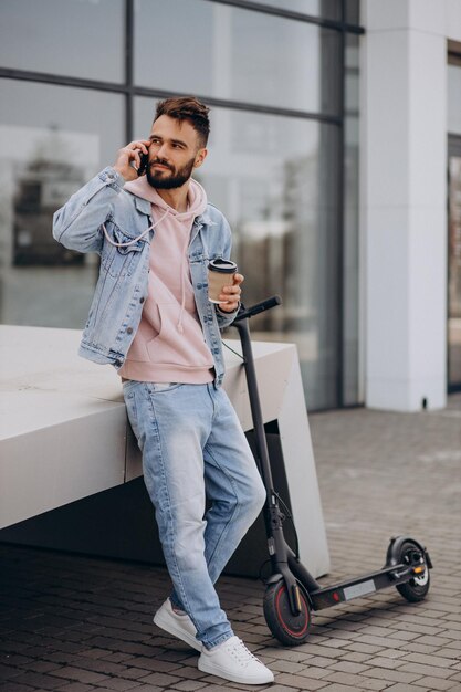 Apuesto joven montando scooter eléctrico usando teléfono y bebiendo café