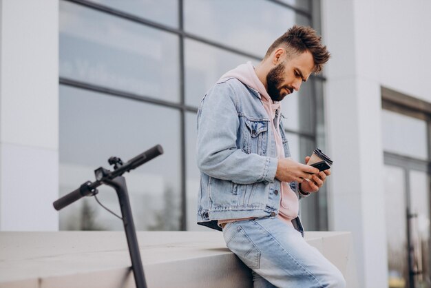 Apuesto joven montando scooter eléctrico usando teléfono y bebiendo café