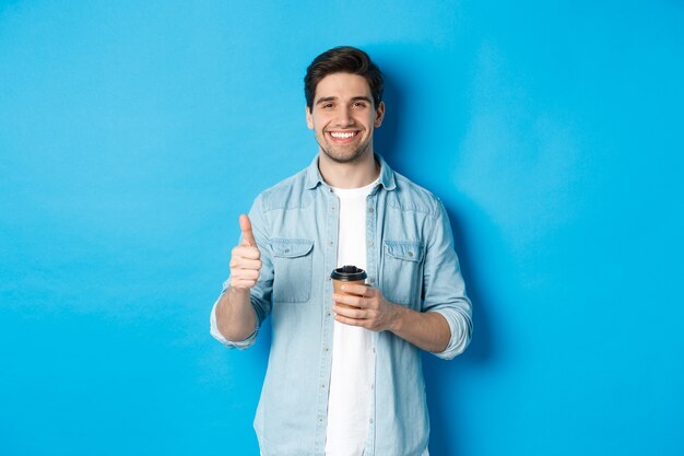 Apuesto joven mostrando el pulgar hacia arriba y tomando café, recomendando café para llevar, de pie sobre fondo azul.