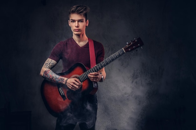 Apuesto joven músico con cabello elegante en una camiseta, sostiene una guitarra en sus manos y toca. Aislado en un fondo oscuro.