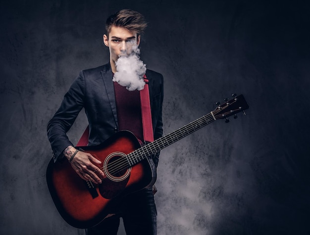 Un apuesto joven músico con cabello elegante y ropa elegante exhala humo mientras toca la guitarra acústica. Aislado en un fondo oscuro.