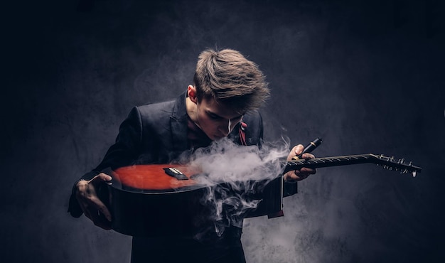 Apuesto joven músico con cabello elegante en ropa elegante exhala humo en su guitarra acústica. Aislado en un fondo oscuro.