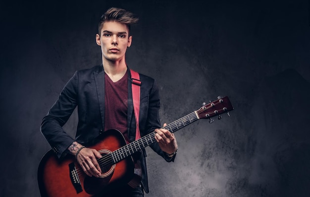 Apuesto joven músico con cabello elegante en ropa elegante con una guitarra en sus manos tocando y posando sobre un fondo oscuro.