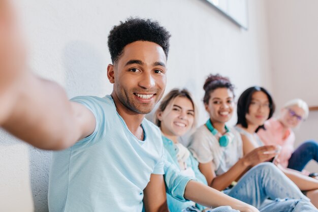 Apuesto joven negro con peinado rizado haciendo selfie con amigos y sonriendo. Retrato interior de estudiantes riendo alegres divirtiéndose después de lección y tomando fotos.