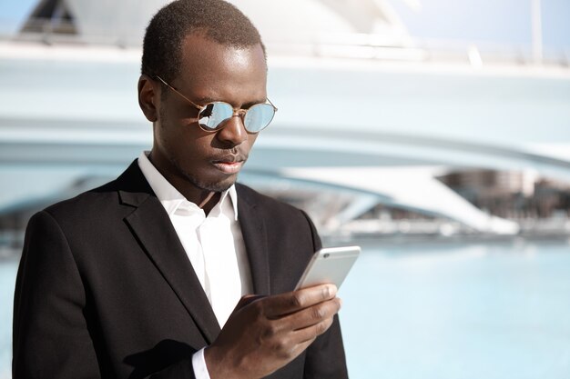 Apuesto joven oficinista afroamericano en elegante traje negro y anteojos de pie en un entorno urbano, mirando concentrado mientras intenta llamar a la cabina usando la aplicación en línea en su teléfono móvil