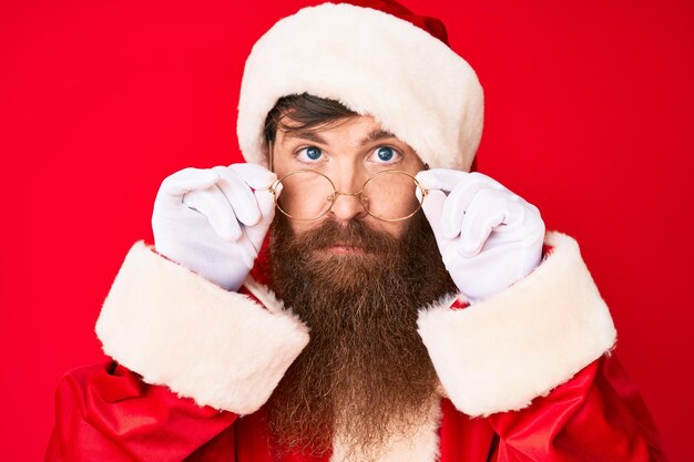 Apuesto joven pelirrojo con barba larga con traje de santa claus y gafas relajado con expresión seria en la cara simple y natural mirando a la cámara