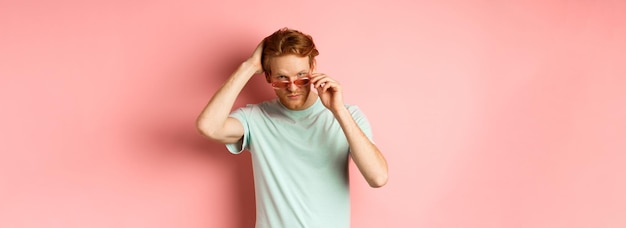 Foto gratuita apuesto joven pelirrojo con gafas de sol cepillando el cabello con la mano y mirando engreído y confiado en c