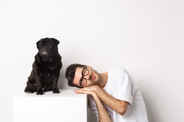 Apuesto joven pone la cabeza cerca de lindo pug negro, sonriendo y mirando el espacio de la copia, fondo blanco.