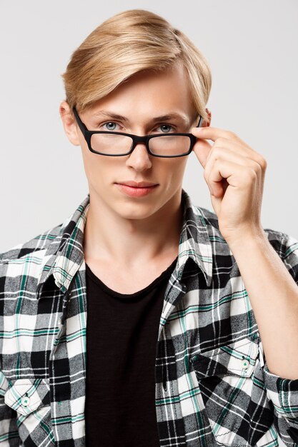 Apuesto joven rubio vistiendo camisa a cuadros casual y gafas en la pared gris