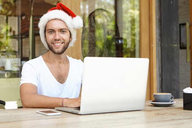 Apuesto joven con sombrero rojo de Santa Claus mirando felizmente a la cámara mientras usa la computadora portátil para el trabajo remoto, pasando las vacaciones de Navidad en un país tropical