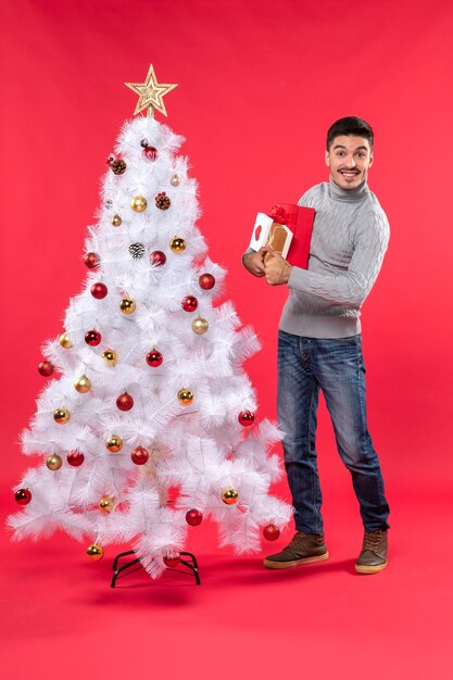 Apuesto joven sonriente de pie cerca del árbol de año nuevo blanco decorado y sosteniendo sus regalos