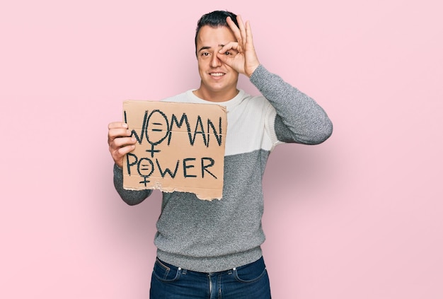 Foto gratuita apuesto joven sosteniendo la pancarta de poder de la mujer sonriendo feliz haciendo el signo de ok con la mano en los ojos mirando a través de los dedos