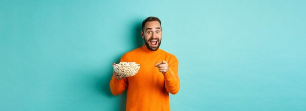 Apuesto joven sosteniendo un tazón de palomitas de maíz sonriendo asombrado y apuntando a la cámara de pie sobre azul