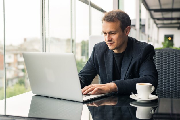 Apuesto joven trabajando en una computadora portátil y sonriendo mientras está sentado en la cafetería en la acera