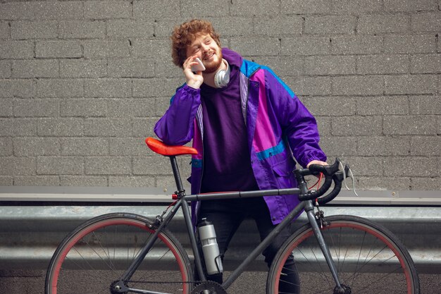 Apuesto joven usando teléfono móvil y auriculares mientras está parado cerca de su bicicleta a su lado