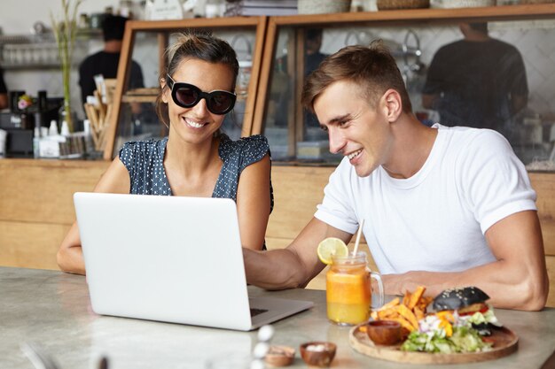 Apuesto joven vistiendo una camiseta blanca que muestra algo en la PC portátil a su atractiva compañera en elegantes gafas de sol durante el almuerzo en el café