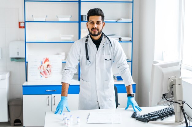 Un apuesto médico indio con una bata médica blanca y un estetoscopio cerca de su lugar de trabajo