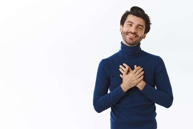 Un apuesto moreno con barba en un suéter de cuello alto presiona las manos contra el corazón y suspira al ver una escena conmovedora mirar con admiración algo adorable sonriendo felizmente recibir un cumplido