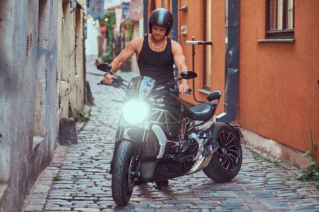 Un apuesto motociclista brutal vestido con una camiseta negra y jeans, parado cerca de una motocicleta, en una estrecha calle vieja de Europa.