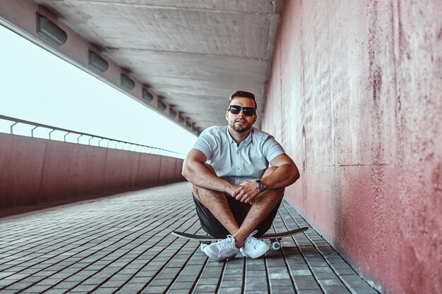 Un apuesto patinador de moda con gafas de sol vestido con una camisa blanca y pantalones cortos sentados en una patineta debajo del puente, mirando una cámara.