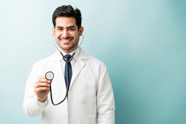 Apuesto profesional médico sonriente examinando con estetoscopio sobre fondo de color