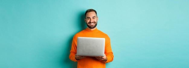 Foto gratuita apuesto trabajador independiente trabajando en una laptop y sonriendo de pie en un suéter naranja sobre un centrico azul claro