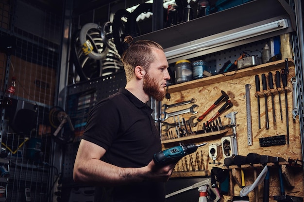 Un apuesto trabajador pelirrojo barbudo con corte de pelo, sosteniendo un destornillador eléctrico, trabajando en un taller de reparación.