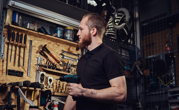 Un apuesto trabajador pelirrojo barbudo con corte de pelo, sosteniendo un destornillador eléctrico, trabajando en un taller de reparación.