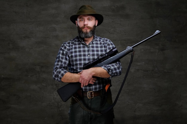 Foto gratuita un apuesto viajero cazador barbudo con una camisa de lana y un sombrero sostiene un rifle con una vista. aislado en un fondo oscuro.