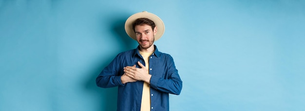 Foto gratuita apuesto viajero con sombrero de paja tomados de la mano en el corazón y sonriendo mostrando gesto de agradecimiento mantener mem