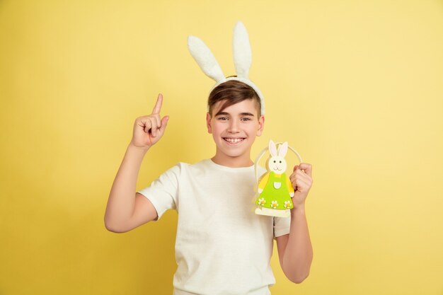 Apuntando hacia Arriba. Decorar. Muchacho caucásico como un conejito de Pascua en el fondo amarillo del estudio. Felices saludos de pascua. Hermoso modelo masculino. Concepto de emociones humanas, expresión facial, vacaciones. Copyspace.