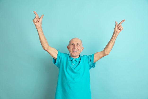 Apuntando hacia Arriba. Retrato de hombre mayor caucásico aislado sobre fondo azul de estudio. Hermoso modelo emocional masculino. Concepto de emociones humanas, expresión facial, ventas, bienestar, publicidad. Copyspace.